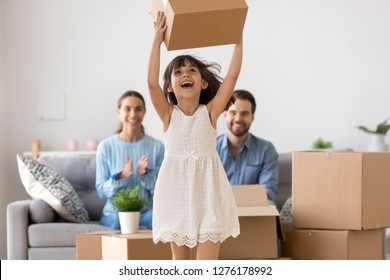 Cute Happy Kid Girl Holding Box Jumping Having Fun Relocating Into New Modern Home Or Apartment With Parents, Excited Child Daughter Playing With Cardboard Box Enjoying Family Moving Day Concept