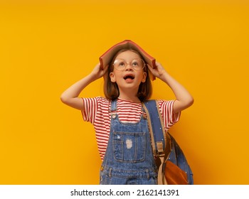 Cute Happy Kid Standing On Bright Yellow Background. Child With Backpack And Book, Little Girl Is Ready To Back To School. 