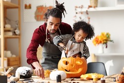 Cute Happy Little African American Boy Carving Halloween Pumpkin With Father At Home, Mixed-race Family Son And Dad Making Jack-o-Lantern Together While Preparing Decorations For Saints Day Party