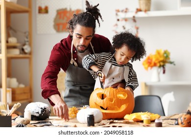 Cute happy little african american boy carving Halloween pumpkin with father at home, mixed-race family son and dad making Jack-o-Lantern together while preparing decorations for Saints Day party
