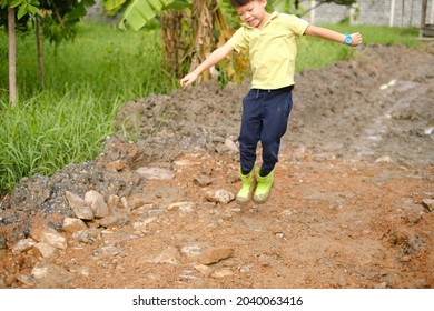 Cute Happy Little Asian 5 Years Old Kindergarten Boy Kid Wearing Green Boots Jump In Muddy Puddles At Home Backyard On Nature, Outdoor Fun In The Rain Weather