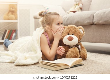 Cute happy little casual girl embracing teddy bear, reading book and sharing secrets with her favorite toy friend. Pretty kid at home, lying on the floor near sofa