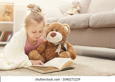 Cute Happy Little Casual Girl Embracing Teddy Bear And Reading Book. Pretty Kid At Home, Lying On The Floor Near Sofa With Her Favorite Toy