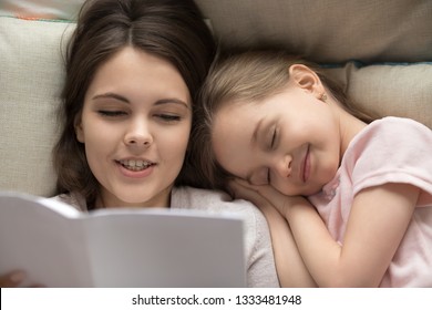 Cute Happy Little Girl Falling Asleep Lying On Mom Shoulder Pillows In Bed While Mum Reading Book Telling Fairy Tale Before Going To Sleep, Mother And Kid Daughter Bedtime Stories Concept, Top View