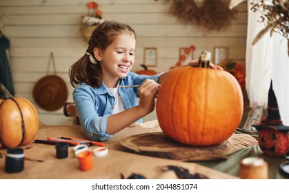 Cute happy little girl preparing for halloween  painting drawing scary face pumpkin while sitting at table in kitchen at home  smiling child making jack  o  lantern  Holiday decoration concept