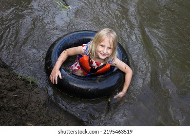 A Cute, Happy, Little Muddy Girl Child Is Swimming In An Inner Tube Raft And Wearing A Life Jacket In A River.