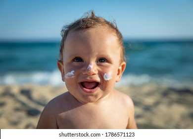Cute Happy Little Toddler Boy Is Smiling In Camera With Protective Sunscreen Or Sunblock Lotion On His Face Applied By His Mother To Take Care Of Skin On Seaside Beach During Family Holidays Vacation.