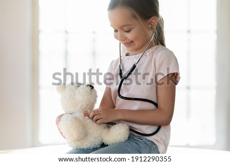 Cute happy small 7s Caucasian girl hold stethoscope act as doctor cure stuffed toy. Smiling little child have fun play hospital listen to fluffy bear heart, dream of future career in medicine.