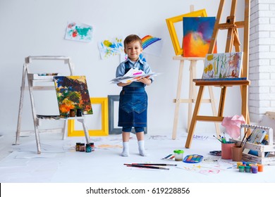 Cute, happy, white boy in blue shirt and jeans smiling and showing his colorful drawings. Little child having fun in artist studio. Concept of early childhood education, happy family, parenting - Powered by Shutterstock