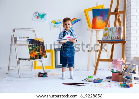 Cute, happy, white boy in blue shirt and jeans smiling and showing his colorful drawings. Little child having fun in artist studio. Concept of early childhood education, happy family, parenting
