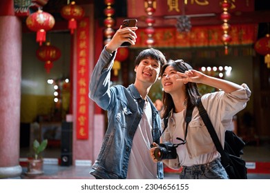 Cute and happy young Asian tourist couple taking their pictures with a smartphone while visiting a Chinese temple together.
