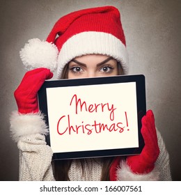 Cute happy young Caucasian woman with Santa hat hiding behind tablet computer with Merry Christmas text message. Christmas and New Year concept.