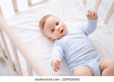 a cute healthy little baby is lying on his back in a crib on white bedding at home in a blue bodysuit. The happy kid looks into the camera, smiles