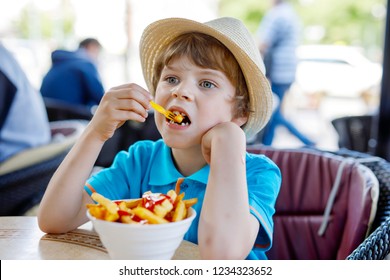 Cute Healthy Preschool Kid Boy Eats French Fries Potatoes With Ketchup Sitting In Cafe Outdoors. Happy Child Eating Unhealthy Food In Restaurant