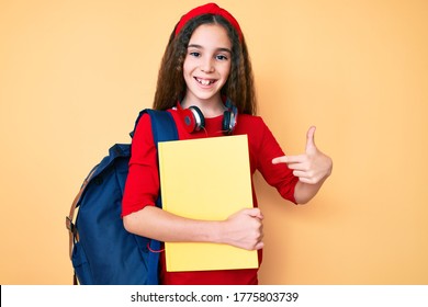 Cute Hispanic Child Girl Wearing Student Backpack And Headphones Holding Book Smiling Happy Pointing With Hand And Finger 