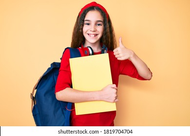 Cute Hispanic Child Girl Wearing Student Backpack And Headphones Holding Book Smiling Happy And Positive, Thumb Up Doing Excellent And Approval Sign 