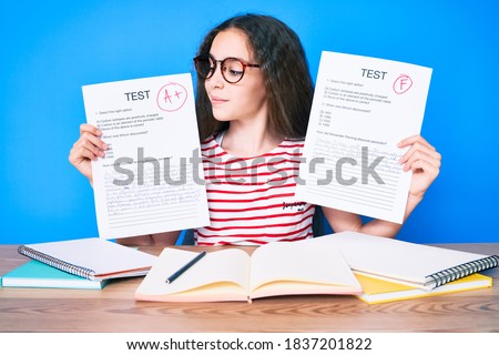 Cute hispanic girl showing failed and passe exam sitting on the table smiling looking to the side and staring away thinking. 