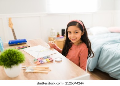 Cute Hispanic Kid Looking At The Camera While Doing A Painting In Her Bedroom Desk For An Art Class Homework 