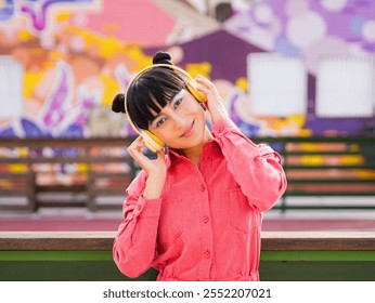 Cute hispanic Woman whit hair buns smile while hear music in yellow headphones, pink jacket and she enjoy. Front Portrait and tilt head whit color city in the background. - Powered by Shutterstock