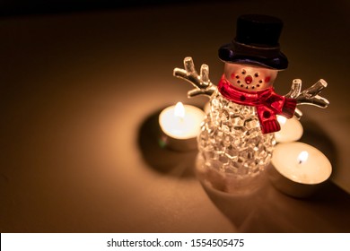 Cute illuminated christmas snowman with a red scarf and a black hat and a smiley face is ready to celebrate advent time and christmas time surrounded by warm shining candle lights at christmas eve