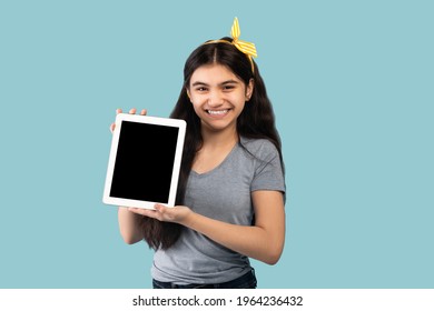Cute Indian adolescent girl holding tablet pc with blank screen on blue studio background, space for app or website design. Smiling teenager showing touch pad with mockup for your ad