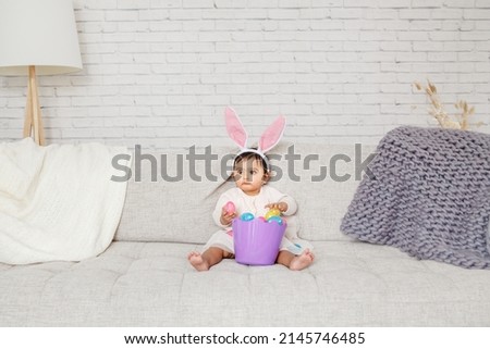 Cute Indian baby girl with pink bunny ears and basket of colorful eggs celebrating Easter holiday.
