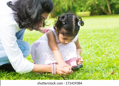Cute Indian girl peeking through magnifying glass with parent on green lawn. Mother and daughter exploring nature at outdoor garden. - Powered by Shutterstock