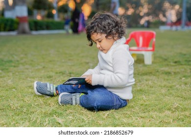 Cute Indian Little Child Face Closeup On Green Grass In Summertime. Funny Little Kid Portrait With Mobile Phone, Smartphone On Nature. Happy Childhood.