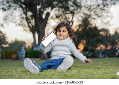 Cute Indian Little Child Face Closeup On Green Grass In Summertime. Funny Little Kid Portrait With Mobile Phone, Smartphone On Nature. Happy Childhood.