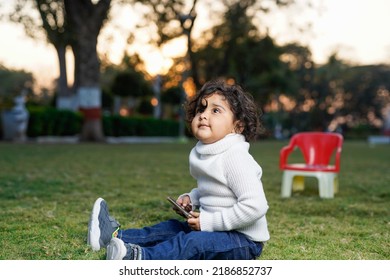 Cute Indian Little Child Face Closeup On Green Grass In Summertime. Funny Little Kid Portrait With Mobile Phone, Smartphone On Nature. Happy Childhood.