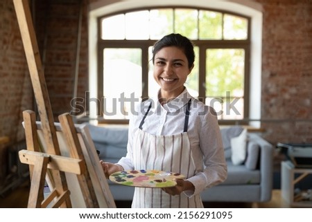 Cute Indian woman painter pose in workshop near easel with canvas holds palette and paintbrush smile look at camera, enjoy study in art-school and creative hobby. Professional artist portrait concept