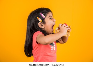 Cute Indian/Asian little girl eating tasty Burger, Sandwich or Pizza in a plate or box. Standing isolated over blue or yellow background.