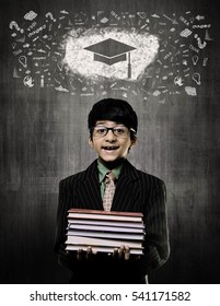 Cute Intelligent Little Boy Holding Books And Wearing Glasses, Smiling While Standing Before A Chalkboard, Graduation Cap Drawn On Board With Some Calkboard Vectors