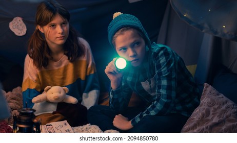 Cute Isolated Caucasian Kids Sitting In A Dark Tent Filled With Cozy Blankets And Pillows, Treasure Map Placed In Front Of Them. Young Girl Scared As The Young Boy Holding A Flashlight Checks Outside.