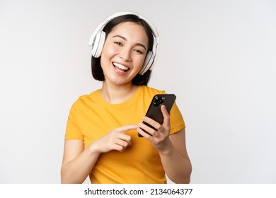 Cute japanese girl in headphones, looking at mobile phone and smiling, using music app on smartphone, standing against white background