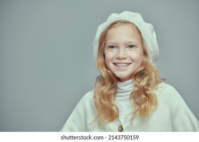 Cute joyful 11 year old girl with blonde hair and freckles poses in a studio in a stylish white beret and sweater. Fashion shot on a gray background. Kid's fashion. 