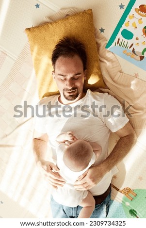 A cute joyful baby and his father are lying on the floor in a bright room. Top view. Happy fatherhood and childhood.