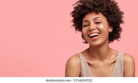 Cute joyful female teenager has broad smile, sparkles on face, prepares to go on disco with friends, being in high spirit, has bare shoulders, stands against pink background, blank space for your text
