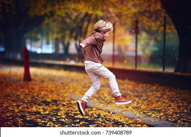 Cute Kid, Boy In Leather Jacket Having Fun At Autumn Street, Jumping And Running Around On Carpet Of Fallen Leaves