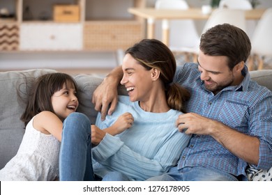 Cute kid daughter and dad tickling mom having fun good time playing together at home, happy parents and little child girl enjoying funny activity and communication, family laughing relaxing on sofa