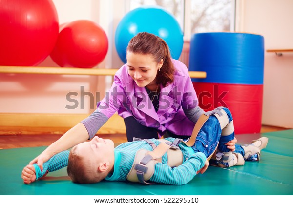 cute kid with disability has\
musculoskeletal therapy by doing exercises in body fixing\
belts