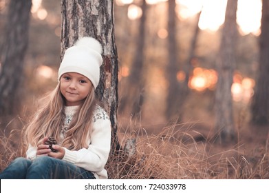 Cute kid girl 4-5 year old wearing knitted sweater and hat sitting under tree in park. Looking at camera. Winter season. Childhood. 
