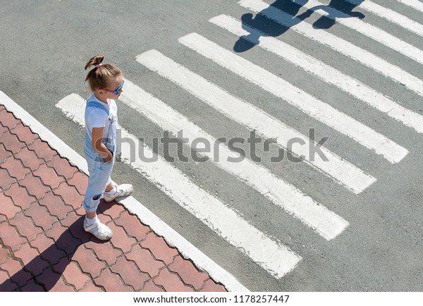 Cute kid girl in glasses and fashion clothes\
standing near the pedestrian crossing on the city street. Shadow\
and silhouette of mother and child crossing the street at zebra\
crosswalk. View from above