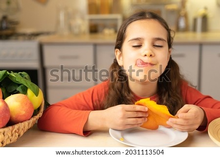 Cute kid girl is happy to eat mangoes. Schoolgirl joy tasty eating and yummy a half of mango on table at home in kitchen
