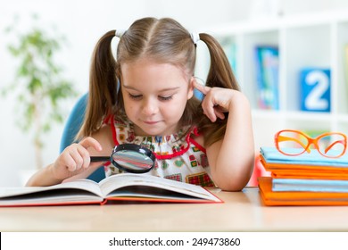 Cute Kid Girl Looks Through A Magnifying Glass With Book Sitting At Table