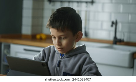 Cute kid sit in the kitchen playing mobile game on digital tablet device. Child surfing internet, play game at home. Children tech addiction concept - Powered by Shutterstock