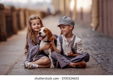 Cute kids boy and girl in retro 
costumes are sitting on the street. They are playing with a beagle dog. Romantic, historical image. Selective focus image.
