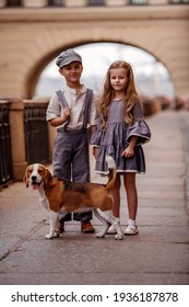 Cute kids boy and girl in retro 
costumes on the street. The girl is holding a leash with a beagle dog. A romantic, historical image. Selective focus of the image.