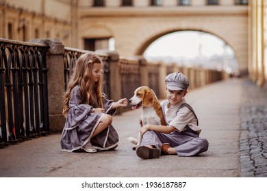 Cute kids boy and girl in vintage costumes are sitting on the street. They are playing with a beagle dog. The boy hugged the dog. The girl feeds her. Romantic, historical image. Selective focus image.