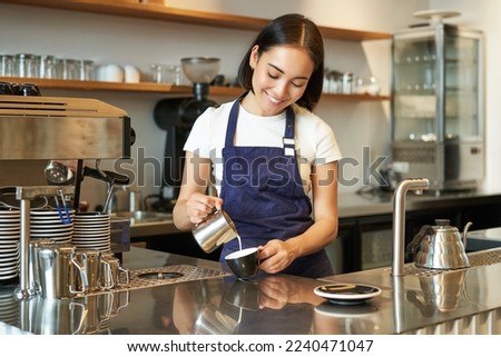 Cute korean barista girl, pouring milk in coffee, prepare cappuccino with latte art, working in cafe behind counter. People and workplace.